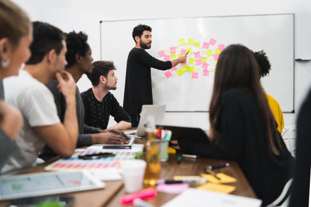 Equipe realizando LNT - Levantamento de necessidades de treinamento em uma reunião colaborativa, com participantes analisando ideias em post-its no quadro branco para identificar demandas de capacitação.