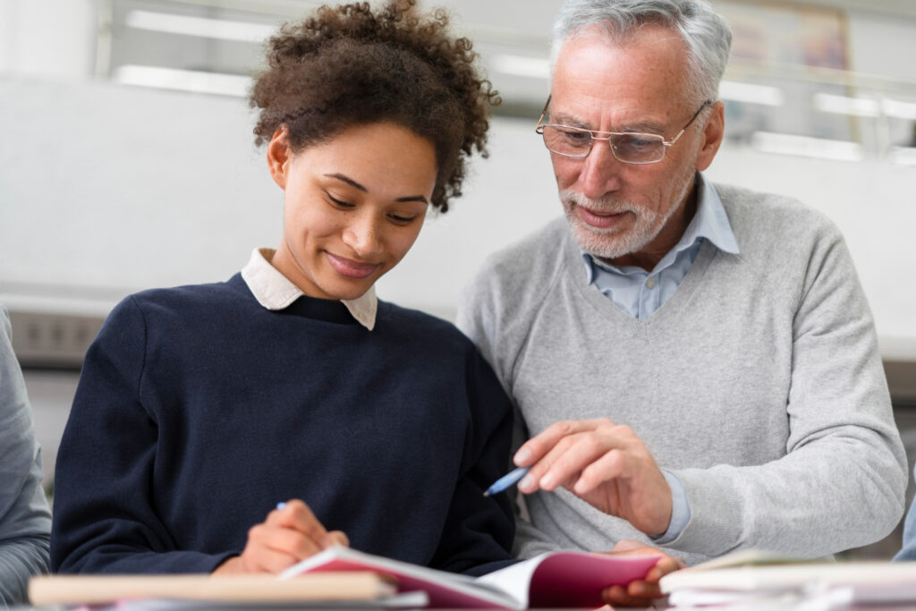 Profissional experiente orientando uma colaboradora durante uma atividade de aprendizagem, enquanto analisam um material de estudo em conjunto. A imagem representa o conceito de lifelong learning, destacando o aprendizado contínuo, a troca de conhecimentos entre gerações e o desenvolvimento de competências no ambiente de trabalho.