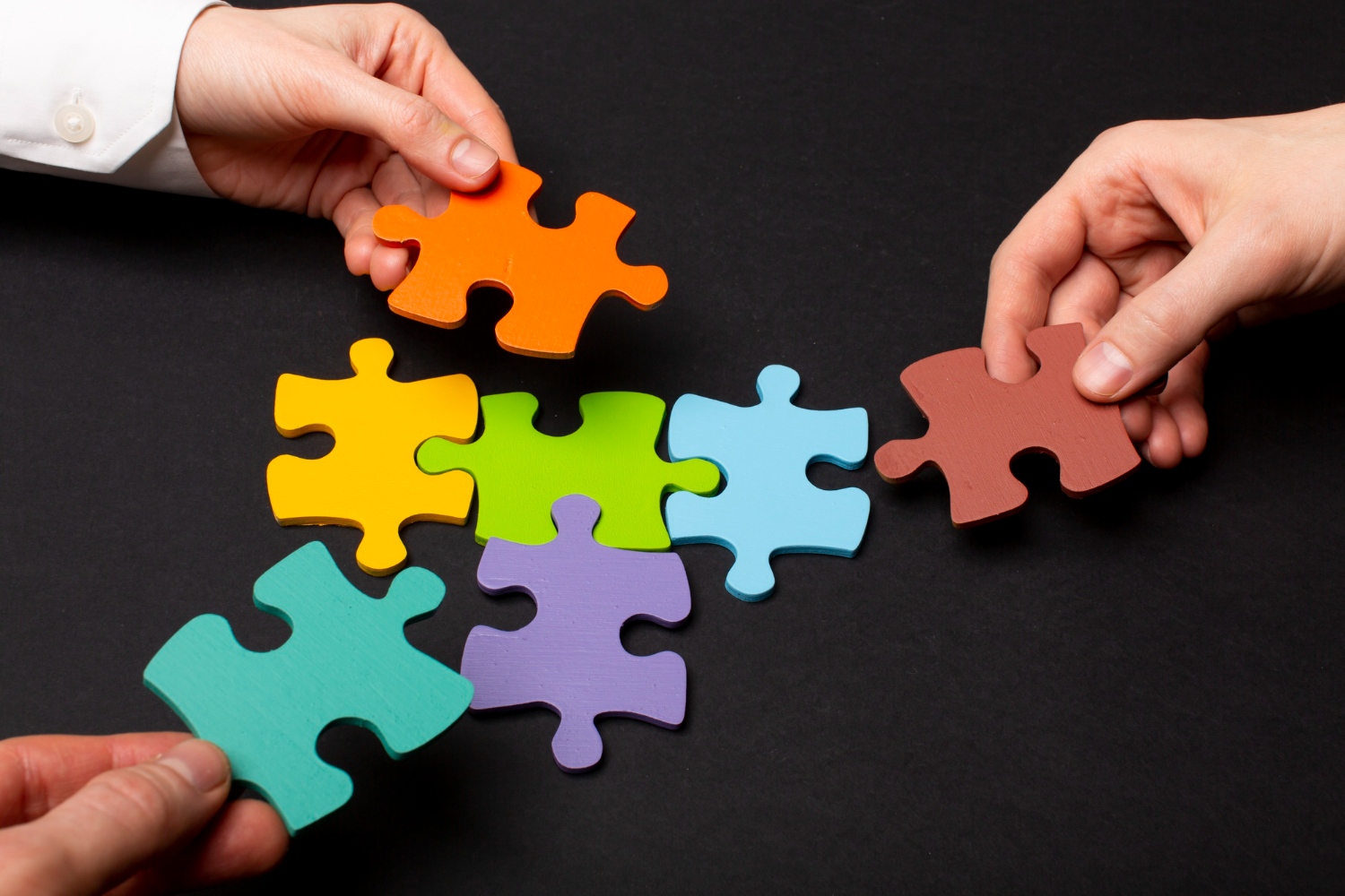 Mãos posicionando peças coloridas de um quebra-cabeça sobre uma superfície escura, simbolizando colaboração, resolução de problemas e construção conjunta de soluções. A imagem representa a adaptabilidade como uma competência essencial para lidar com mudanças, integrar diferentes perspectivas e responder aos desafios do ambiente de trabalho.